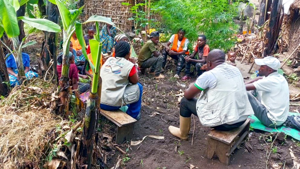 A group of people seated outdoors in a garden-like setting, engaged in discussion, surrounded by greenery and banana plants.