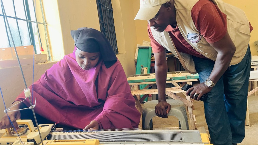 A woman in a purple garment working on a knitting machine, guided by a man in a beige vest and cap. They are in a well-lit room with wooden furniture.